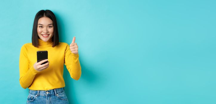 E-commerce and online shopping concept. Satisfied asian female client showing thumbs up and using smartphone, smiling pleased at camera, blue background.