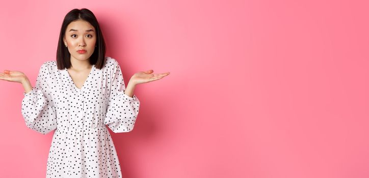 Indecisive cute asian woman shrugging, holding hands spread sideways on copy spaces, dont know, having doubts, standing over pink background.