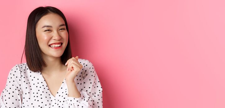 Beauty and lifestyle concept. Close-up of happy asian female laughing, looking happy and showing genuine emotions, standing over pink background.