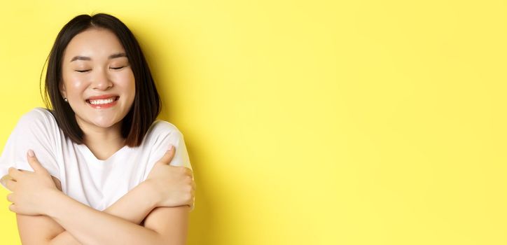 Close up of romantic asian girl hugging herself and dreaming, close eyes and smile while imaging something tender, standing over yellow background.