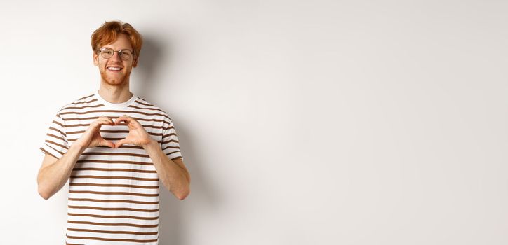 Valentines day. Happy boyfriend with red hair, smiling and showing heart gesture, I love you, standing over white background.