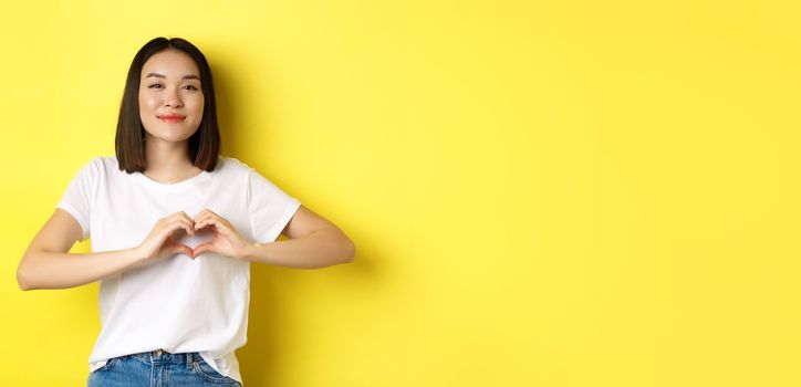 Beautiful asian woman showing I love you heart gesture, smiling at camera, standing against yellow background. Concept of valentines day and romance.