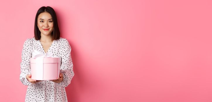 Beautiful asian woman wishing happy holidays, giving you gift in cute box, standing against pink background.