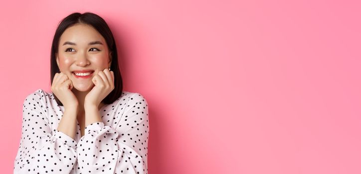 Beauty and skin care concept. Close-up of beautiful asian woman with clean face, blushing and looking dreamy left, admiring promo offer, standing over pink background.