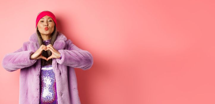 Romance and valentines day. Happy asian senior woman showing heart sign, I love you gesture, pucker lips for kiss, standing over pink background.