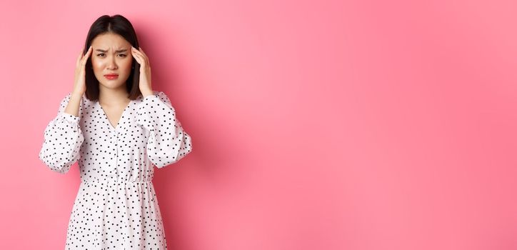 Image of upset asian woman having headache, feeling unwell or dizzy, close eyes and massaging head, suffering migraine, standing over pink background.