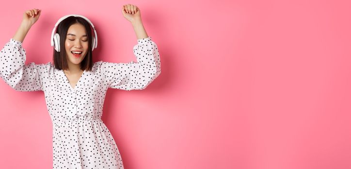 Happy young asian woman dancing and having fun, listening music in headphones, standing over pink background.
