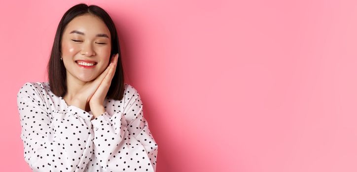 Beauty and lifestyle concept. Close-up of beautiful and dreamy asian woman sleeping on her hands, close eyes and smiling, daydreaming on pink background.