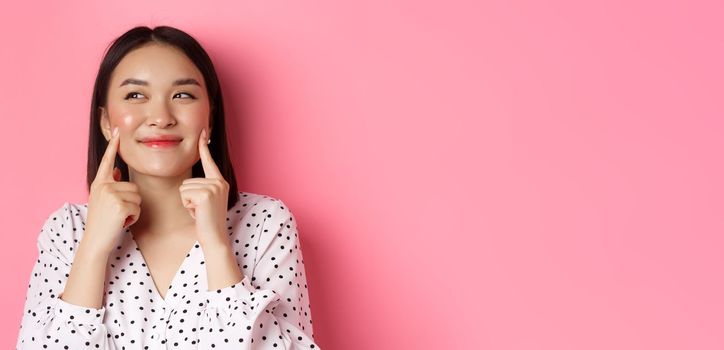 Beauty and skin care concept. Close-up of beautiful asian woman poking cheeks and smiling satisfied, standing over pink background.