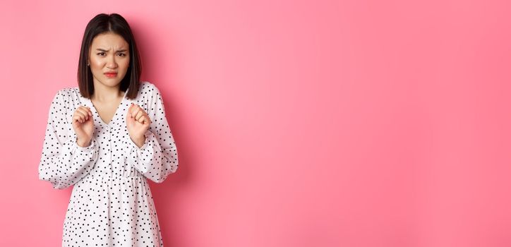 Disgusted asian woman staring with aversion and dislike, frowning and grimacing dissatisfied, standing in dress against pink background.