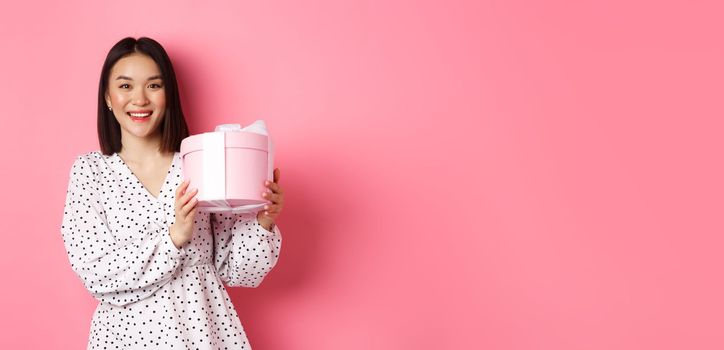 Romantic asian woman in cute dress holding box with gift, smiling happy at camera, standing with present over pink background.