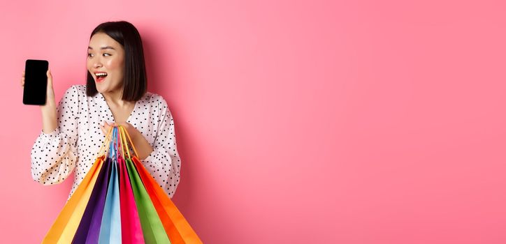 Attractive asian woman showing smartphone app and shopping bags, buying online via application, standing over pink background.