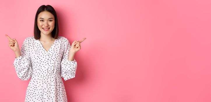 Beautiful asian woman making choice on shopping, pointing fingers sideways and showing variants, smiling at camera, standing over pink background.