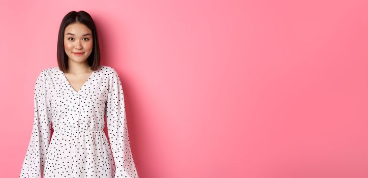 Beautiful asian lady smiling at camera, wearing cute romantic dress, standing over pink background. Copy space