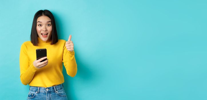 E-commerce and online shopping concept. Excited and amazed asian woman showing thumbs up after using smartphone app, recommend device, standing over blue background.