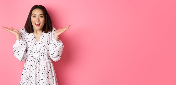 Image of surprised korean girl in dress, female model staring at camera and gasping amazd, standing over pink background.