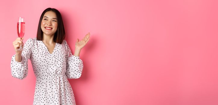 Beautiful asian woman, raising glass of champagne and smiling happy, drinking wine and celebrating, standing over pink background.