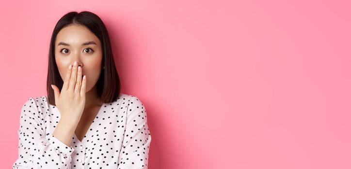 Image of shocked asian girl gossiping, gasping and cover mouth, stare at camera with complete disbelief, standing over pink background.