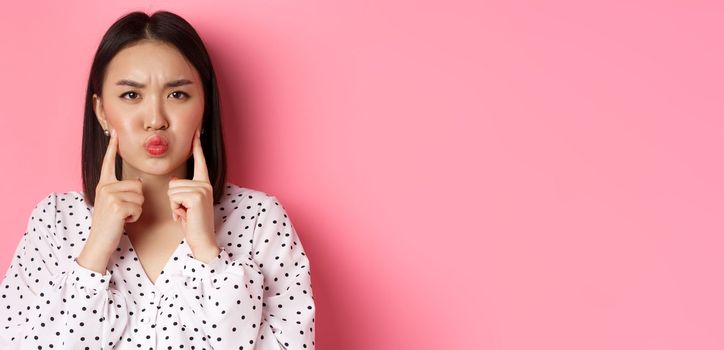 Beauty and lifestyle concept. Cute asian woman making grumpy face, poking cheeks and grimacing, standing over pink background.