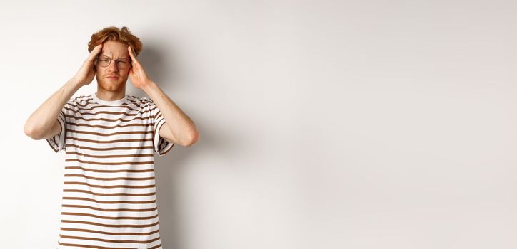 Image of young man with red hair and glasses touching head, frowning from painful migraine, having headache, standing over white background.