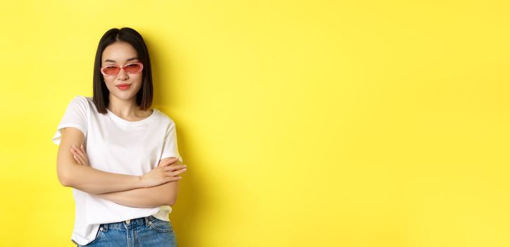 Fashion and lifestyle concept. Sassy and confident asian woman in trendy sunglasses looking self-assured at camera, standing over yellow background.