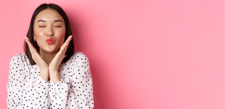 Beauty and skin care concept. Close-up of beautiful asian girl close eyes, pucker lips for kisses, showing cute perfect face, standing over pink background.