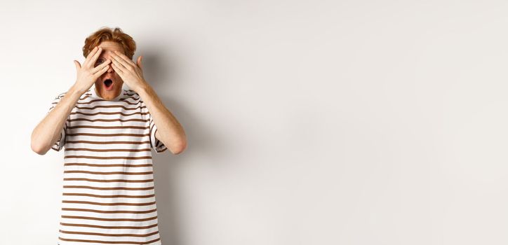 Shocked and embarrassed redhead guy covering eyes, peeking through fingers and gasping, standing over white background.