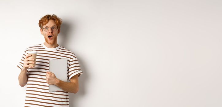 Amazed redhead man talking with coworkers on coffee break, holding cup and laptop, staring surprised at camera, white background.