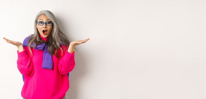 Fashionable asian senior woman in sunglasses gasping amazed, spread hands sideways and staring surprised at camera, white background.