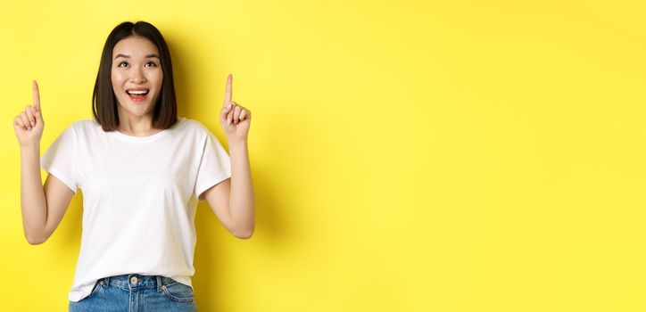 Beauty and fashion concept. Beautiful asian woman in white t-shirt pointing fingers up, standing over yellow background.