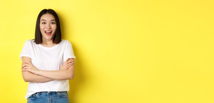 Beauty and fashion concept. Happy asian woman in white t-shirt, cross arms on chest, looking amazed at camera, standing over yellow background.