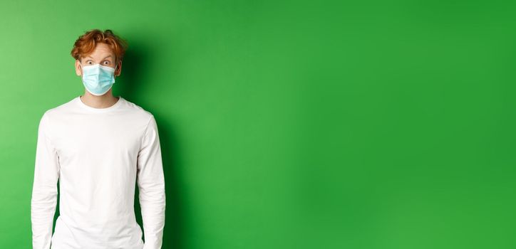 Covid-19, social distancing and lifestyle concept. Young redhead man wearing face mask during coronavirus pandemic, looking surprised at camera, green background.