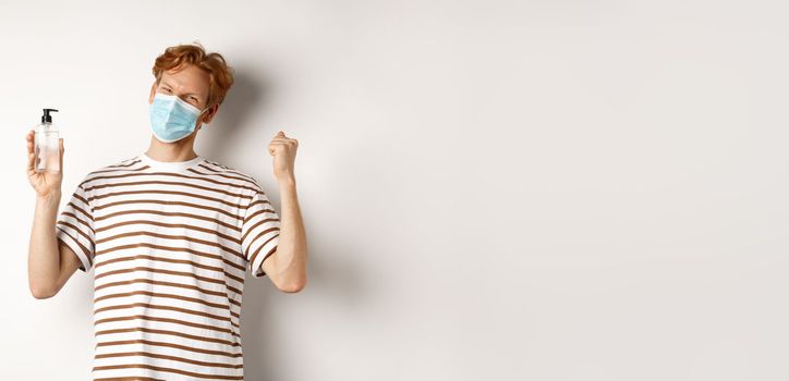 Covid-19, health and lifestyle concept. Handsome young man with red hair wear face mask and celebrating, showing hand sanitizer and looking cheerful, white background.