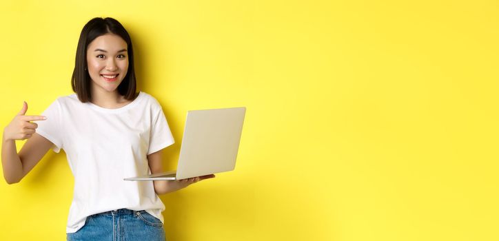 Smiling asian woman pointing finger at her laptop, showing something online, standing over yellow background.