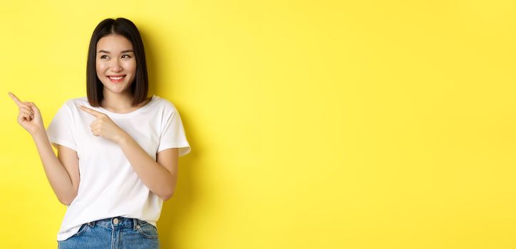 Beauty and fashion concept. Beautiful asian woman in white t-shirt pointing fingers left, standing over yellow background.