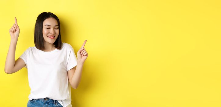 Happy asian woman dancing and having fun, posing in white t-shirt against yellow background.