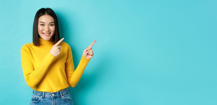 Shopping concept. Attractive asian girl in trendy sweater showing advertisement, pointing fingers right and smiling, recommend promotion deal, blue background.