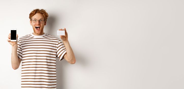 Shopping and finance concept. Young man showing blank mobile screen and plastic credit card, smiling at camera, white background.