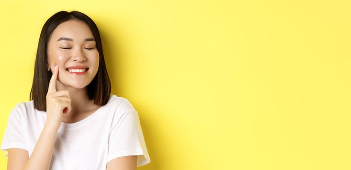 Beauty and skincare. Close up of young asian woman with short dark hair, healthy glowing skin, smiling and touching dimples on cheeks, standing over yellow background.