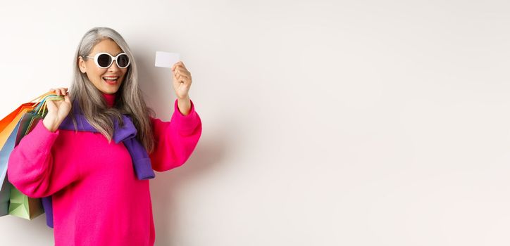 Fashionable asian old lady going shopping, holding paper bags and plastic credit card, wearing stylish sunglasses, standing over white background.