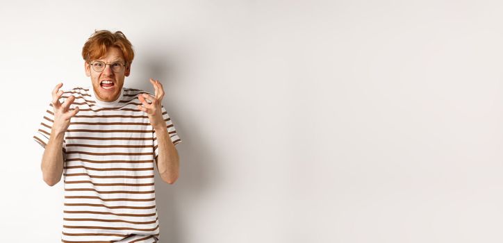 Portrait of redhead male student looking annoyed and pissed-off, shaking hands and staring angry at camera, shouting at someone, standing over white background.