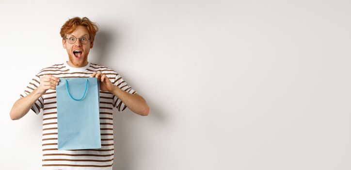 Holidays and celebration concept. Surprised redhead man receiving present inside shopping bag, looking amazed and thankful at camera, white background.