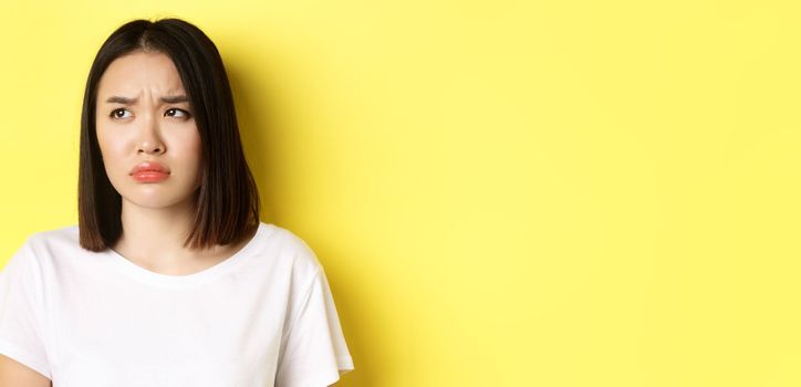 Sad and jealous asian girl sulking, frowning and looking left with upset face, standing over yellow background.