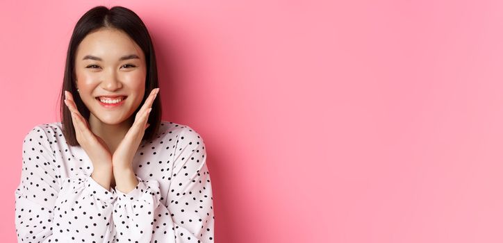 Beauty and skin care concept. Close-up of cute asian woman showing clean perfect face and smiling, looking happy at camera, standing over pink background.
