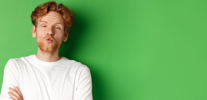 Emotions and fashion concept. Silly redhead guy with beard pucker lips and leaning for kiss, standing over green background.