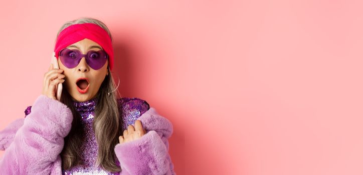 Fashion. Close-up of stylish asian old woman hear shocking news on smartphone, gasping and staring amazed at camera, wearing trendy sunglasses, pink background.