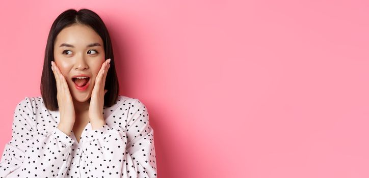 Close-up of beautiful asian woman looking surprised and excited, hear amazing news, looking left and rejoicing, standing against pink background.