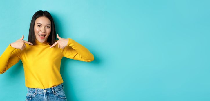 Beauty and fashion concept. Confident and sassy asian woman pointing at herself, smiling cheeky at camera, being professional, standing over blue background.