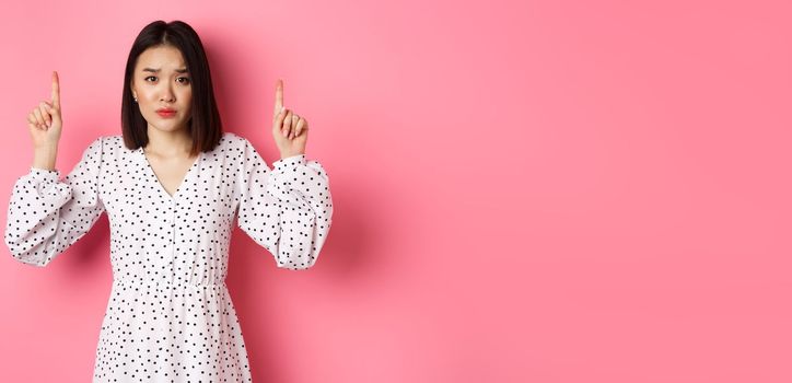 Upset asian girl pointing fingers up, showing promo banner, staring at camera with disappointed gloomy look, standing in dress over pink background.