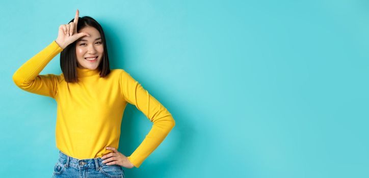 Sassy asian girl mocking lost team, showing loser sign on forehead and smiling pleased, being a winner, standing over blue background.